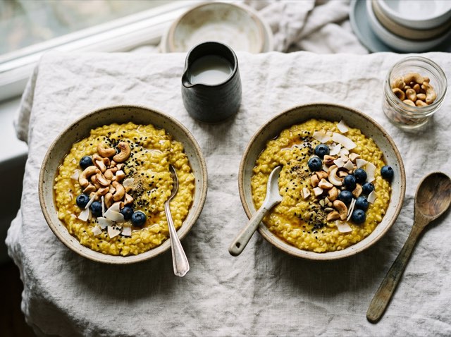 Two bowls of golden turmeric oatmeal topped with toasted chopped cashews on a bright linen surface.