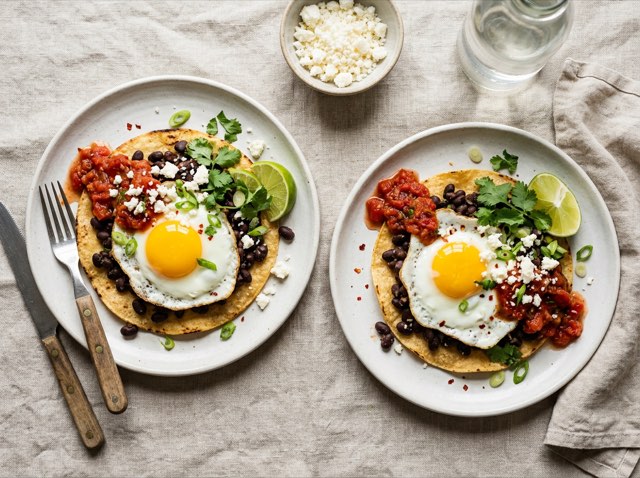 Two sunny-side fried eggs over seasoned black beans topped with red salsa and crumbled feta cheese on a ceramic plate.
